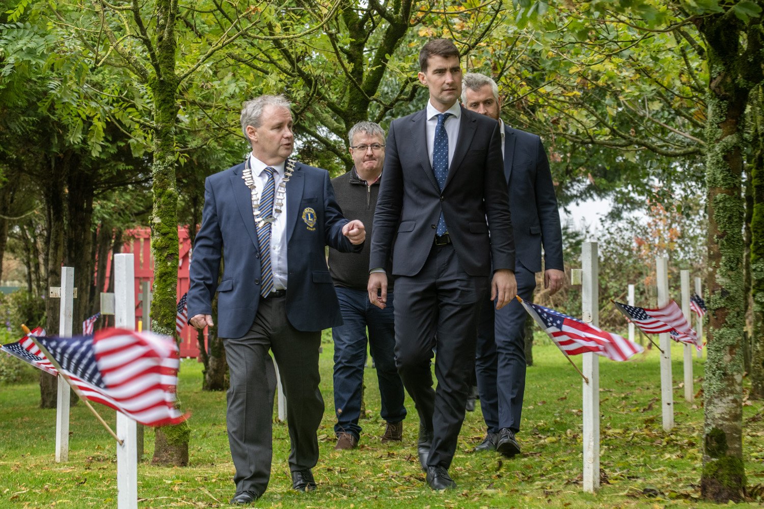 Four men walking between white crosses decorated with small American flags, one wearing a Lions Club chain of office