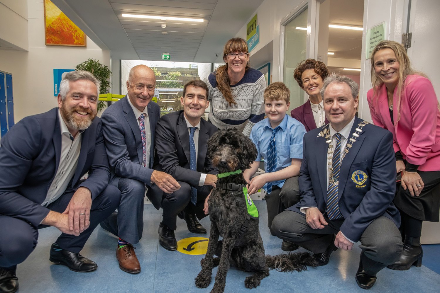 Lions Club president in his chain of office, a schoolboy, several adults and a black dog in a green harness kneeling together for a group photo