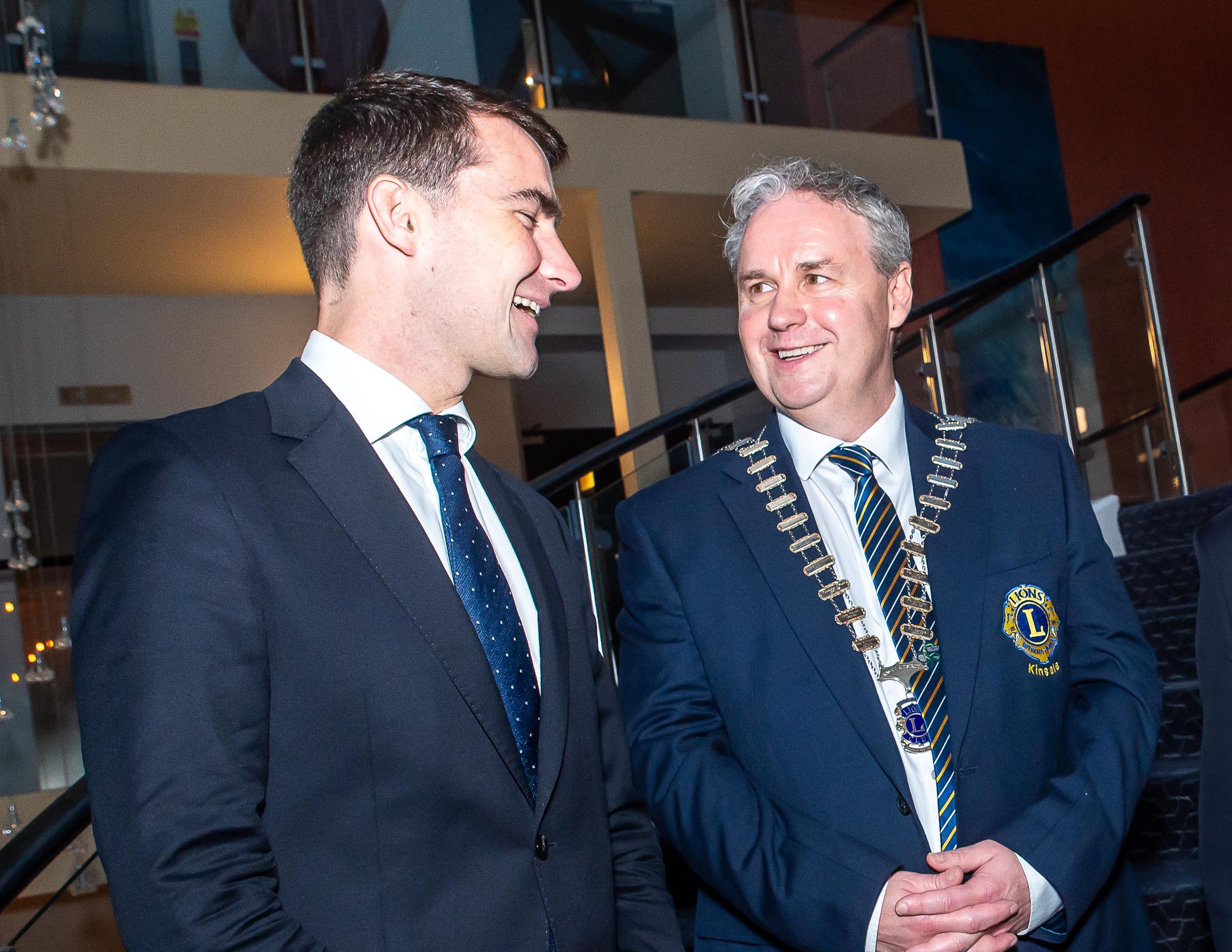 Two men in formal attire standing in conversation. One wears a badge and chain, indicating involvement in an organization. They appear to be in an indoor setting with stairs in the background.