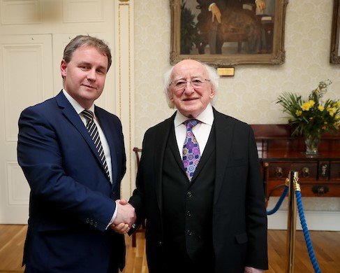 Two men in formal attire shaking hands in a room with elegant decor, including a framed painting and floral arrangement on a wooden console table.