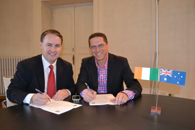 Two men in suits sitting at a table signing documents, with Irish and Australian flag on the table.