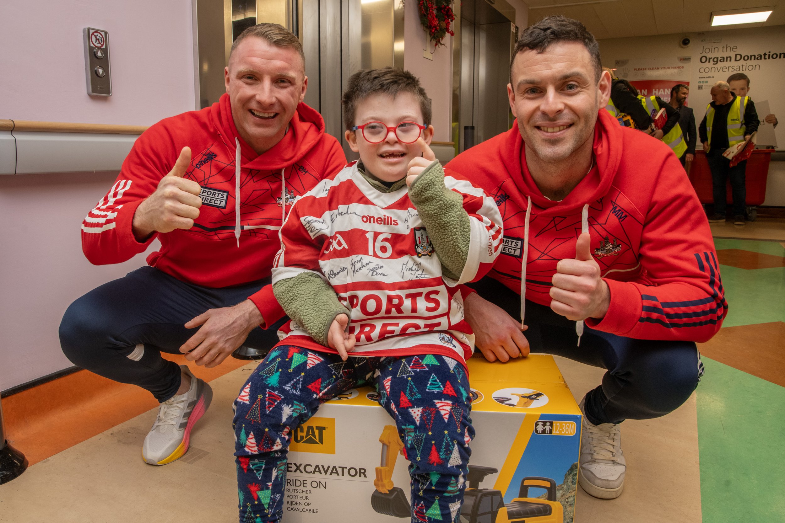 Two men in red tracksuits giving a thumbs-up beside a smiling boy in a red-and-white jersey sitting on a boxed toy excavator in a hospital corridor