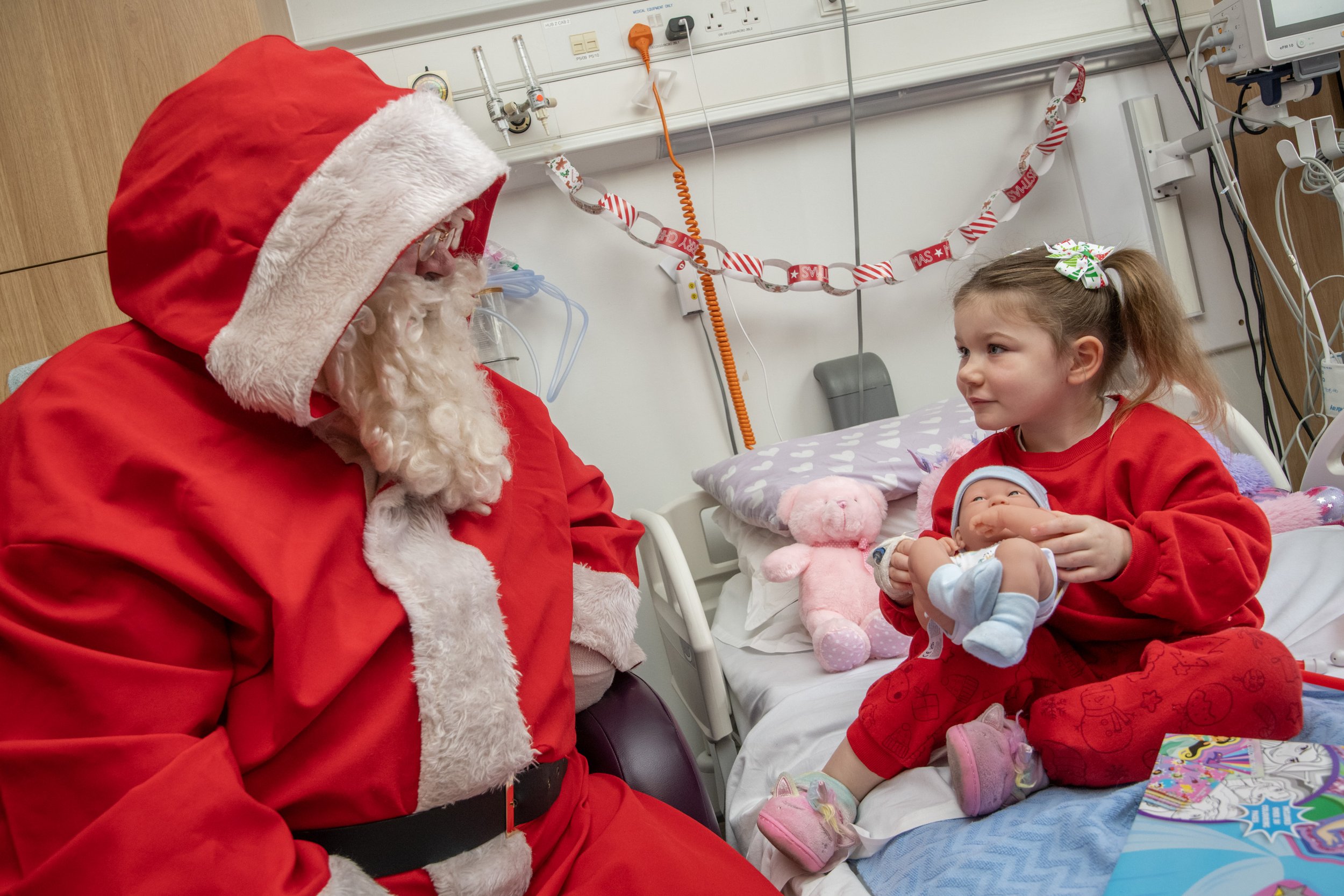 Santa Claus sitting beside a young girl holding a doll on a hospital bed