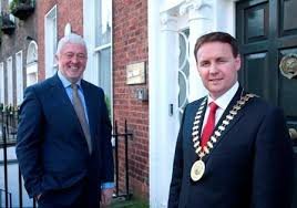 Two men in suits, one wearing a chain of office medallion, standing outside a brick building with a black door.