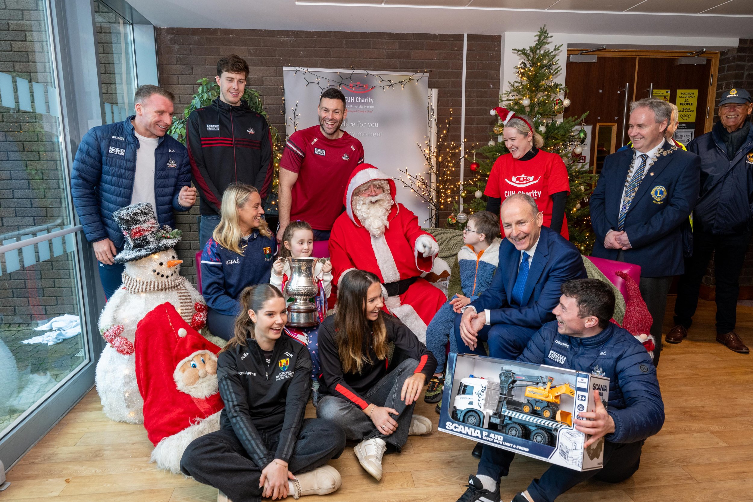 Santa Claus with children, hospital staff, sports players and Lions Club members around a Christmas tree, with a boxed toy truck in the foreground