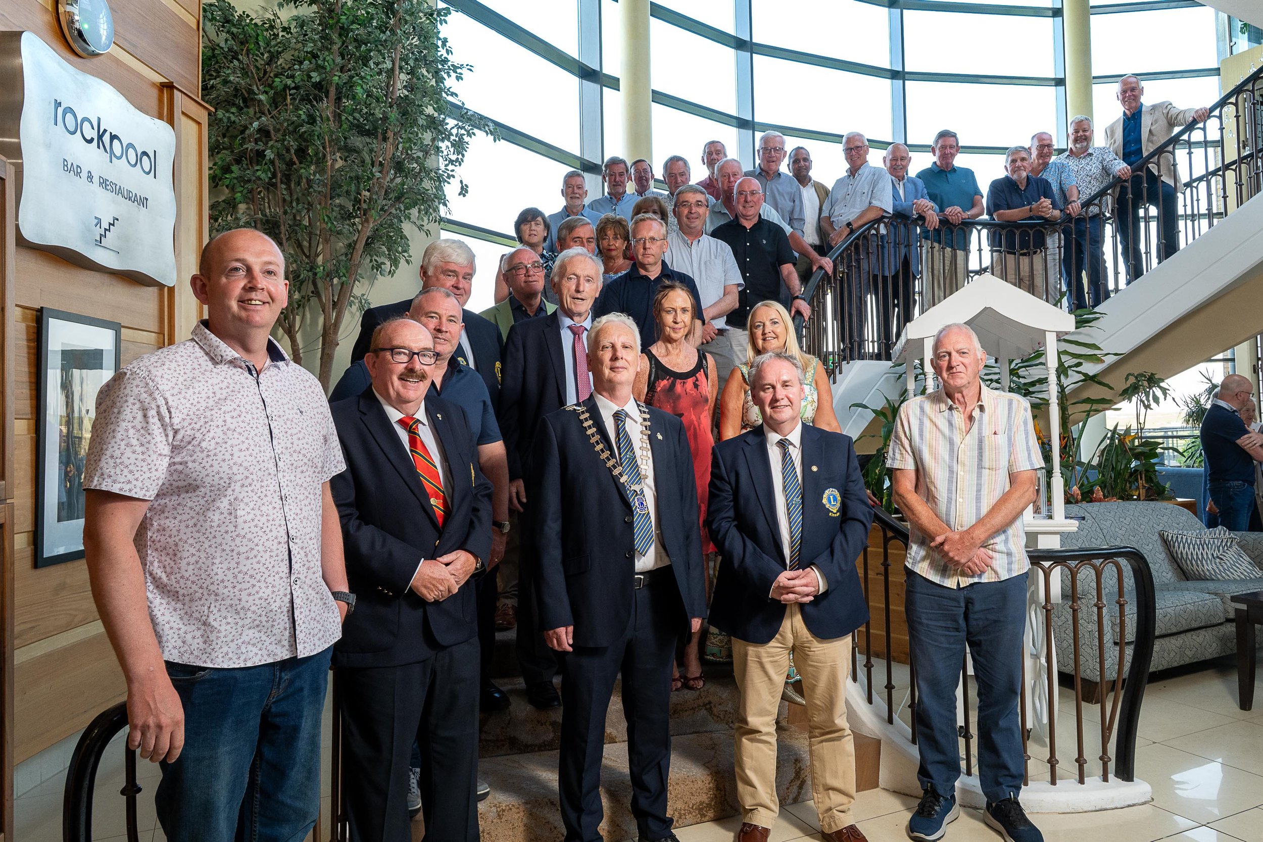 A large group of men and women gathered on a staircase in a hotel foyer, with the Kinsale Lions Club president in the front row wearing his chain of office