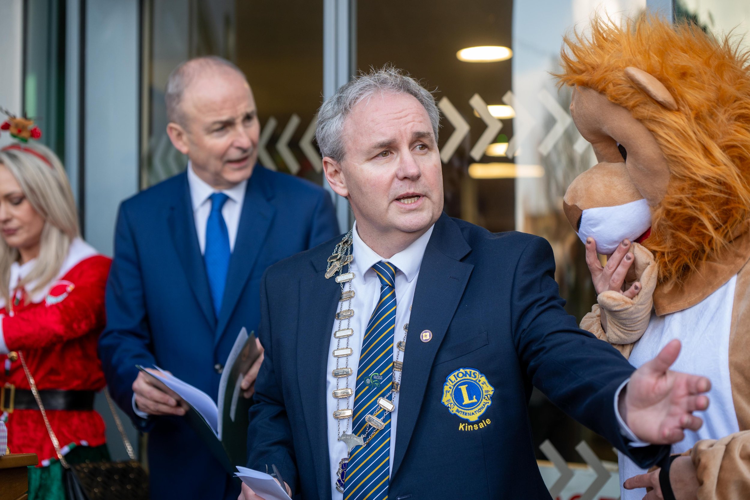 Kinsale Lions Club president in his chain of office speaking beside a man in a suit and a person in a lion mascot costume