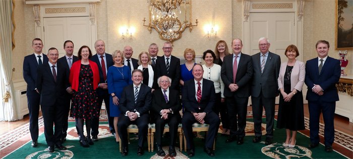 Group portrait of about twenty people in a formal reception room beneath a chandelier, with three men seated at the front