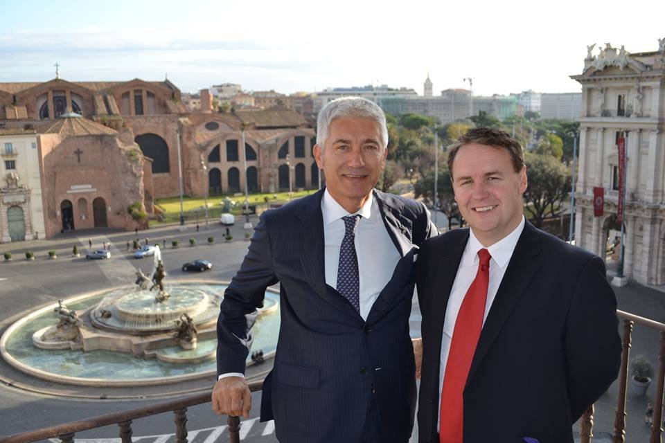Two men in suits standing on a balcony overlooking Piazza della Repubblica with Fountain of the Naiads in Rome.